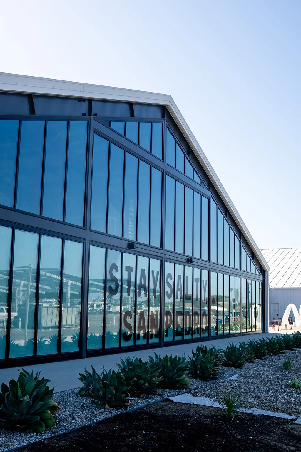 Glass-walled pavilion at West Harbor reflecting the harbor, with 'Stay Salty San Pedro' lettering on the glass